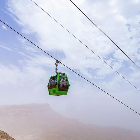 Tolip Resort El Galala Majestic Ain Sukhna Ngoại thất bức ảnh The photo shows a green cable car suspended in the air, traveling along overhead cables. The backdrop features a hazy landscape, likely mountainous or elevated terrain, with a cloudy sky. The scene suggests a scenic transportation method, possibly us