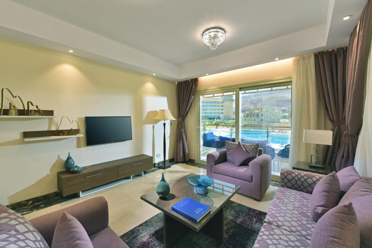 Tolip Resort El Galala Majestic Ain Sukhna Ngoại thất bức ảnh The photo shows a modern living room that features a stylish and cozy design. There are two purple sofas with decorative cushions arranged around a coffee table. The coffee table has a blue book and a decorative item on it. A flat-screen TV is mounte