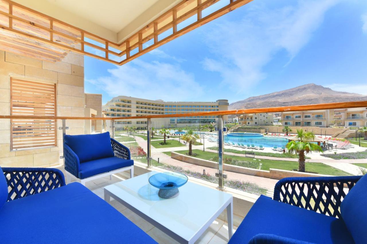 Tolip Resort El Galala Majestic Ain Sukhna Ngoại thất bức ảnh The photo showcases a balcony view featuring two blue chairs and a table with a decorative bowl. The balcony overlooks a landscape that includes a swimming pool and manicured lawns. In the background, there are multiple buildings and palm trees, all 