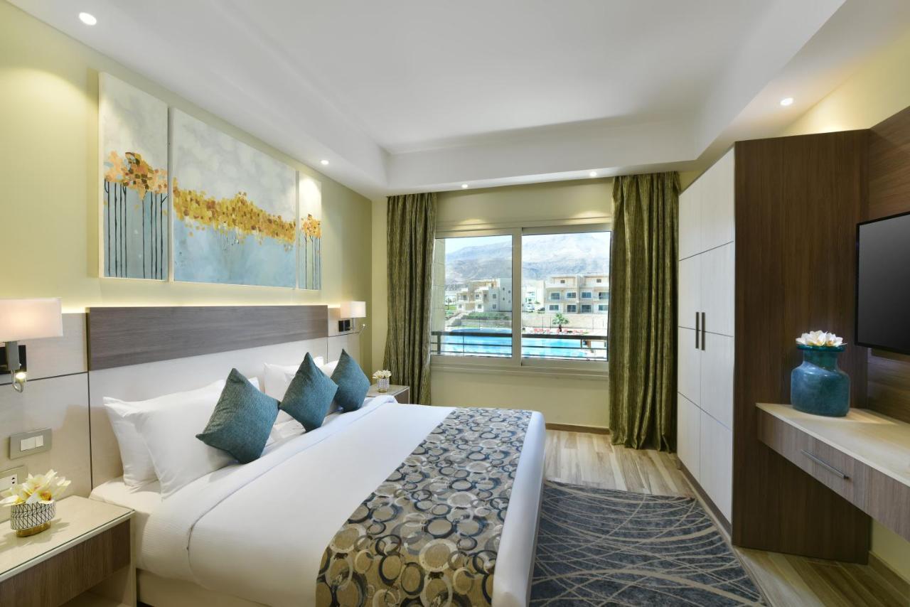Tolip Resort El Galala Majestic Ain Sukhna Ngoại thất bức ảnh The photo shows a modern hotel room. It features a large bed with white bedding and several decorative pillows in shades of teal. Above the bed, there are two pieces of wall art with a landscape theme. The room has a large window with green curtains,