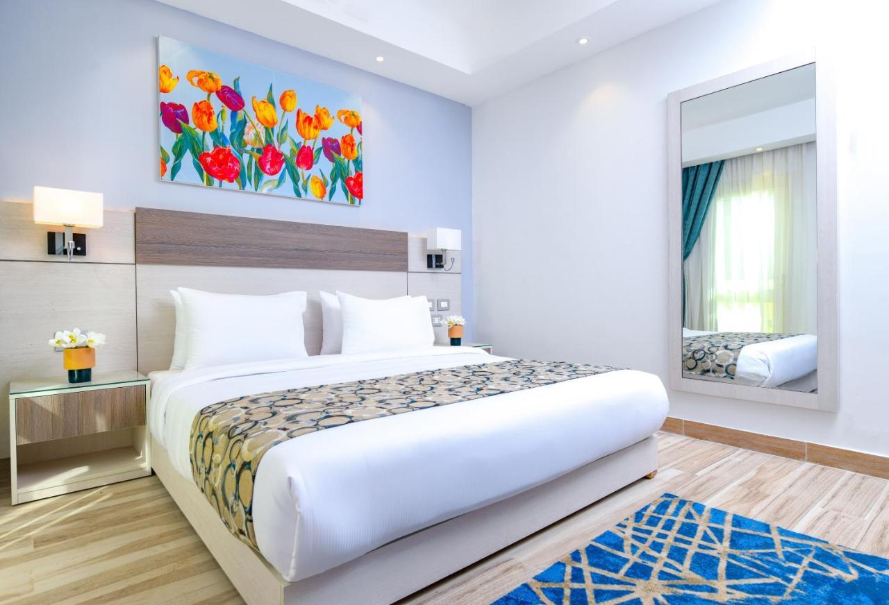 Tolip Resort El Galala Majestic Ain Sukhna Ngoại thất bức ảnh The photo shows a modern hotel bedroom. It features a large bed with white linens and a decorative throw blanket. The room has light wooden flooring and is bright, with ample natural light coming from a window. There is a colorful painting of flowers