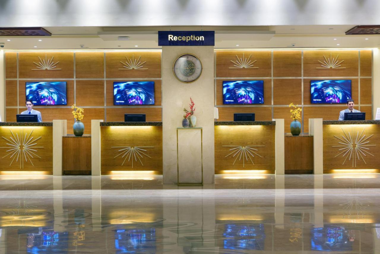 Tolip Resort El Galala Majestic Ain Sukhna Ngoại thất bức ảnh The image shows a hotel reception area. There are several reception desks lined up against a wall with decorative paneling. Each desk has a staff member, and above the desks, there are screens displaying information. A clock is visible on the wall, a