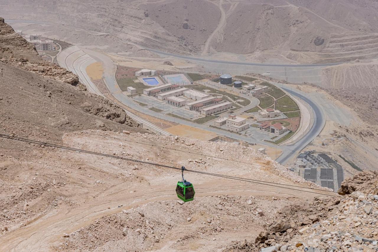 Tolip Resort El Galala Majestic Ain Sukhna Ngoại thất bức ảnh The photo shows a mountainous landscape with a steep, dry terrain. In the foreground, there is a green cable car or gondola traveling along a cable. Below, a complex of buildings and roads can be seen, likely part of a resort or recreational area. Th