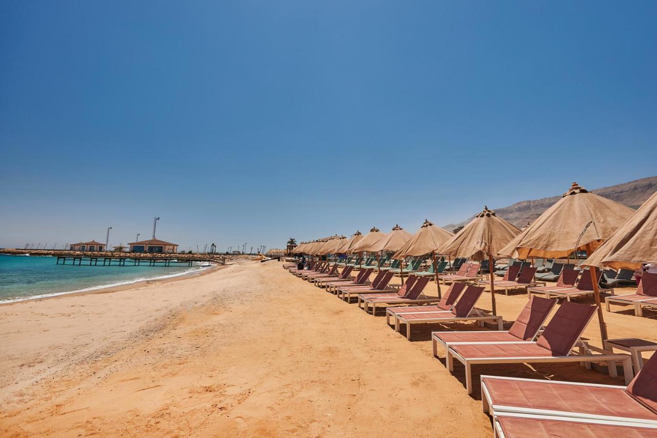 Tolip Resort El Galala Majestic Ain Sukhna Ngoại thất bức ảnh The photo shows a scenic beach with a stretch of golden sand under a clear blue sky. There are several beach loungers arranged in rows, each accompanied by thatched umbrellas providing shade. In the background, there is a wooden pier extending into t