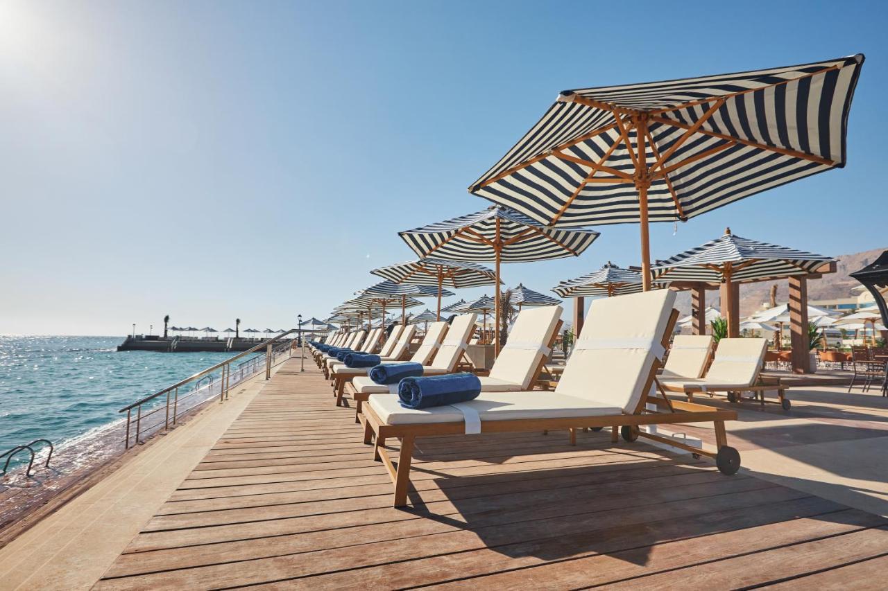 Tolip Resort El Galala Majestic Ain Sukhna Ngoại thất bức ảnh The photo depicts a serene beachside setting featuring a row of sun loungers arranged on a wooden deck. Each lounger is equipped with a folded towel and shaded by striped umbrellas. In the background, the water sparkles under bright sunlight, and the