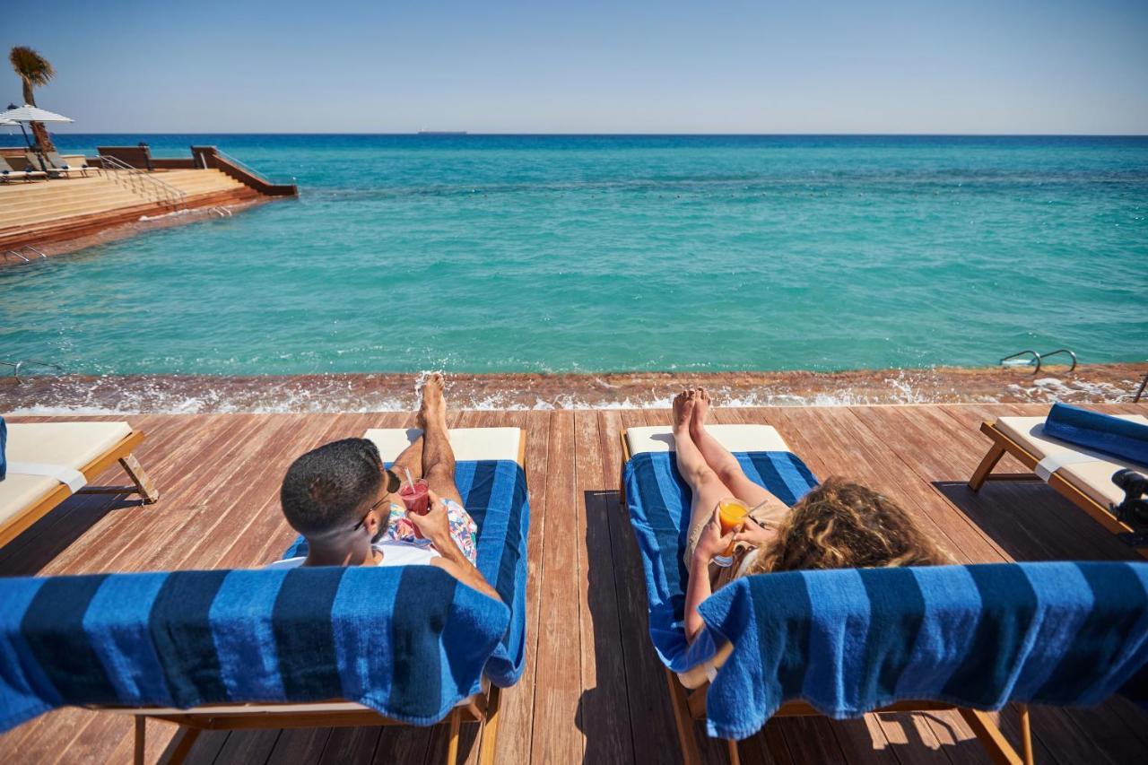 Tolip Resort El Galala Majestic Ain Sukhna Ngoại thất bức ảnh The photo depicts two people relaxing on lounge chairs by the waterfront. They are positioned on a wooden deck with a view of the clear, turquoise water. Both individuals are reclining, enjoying the sun, and each has a drink in hand. The scene convey