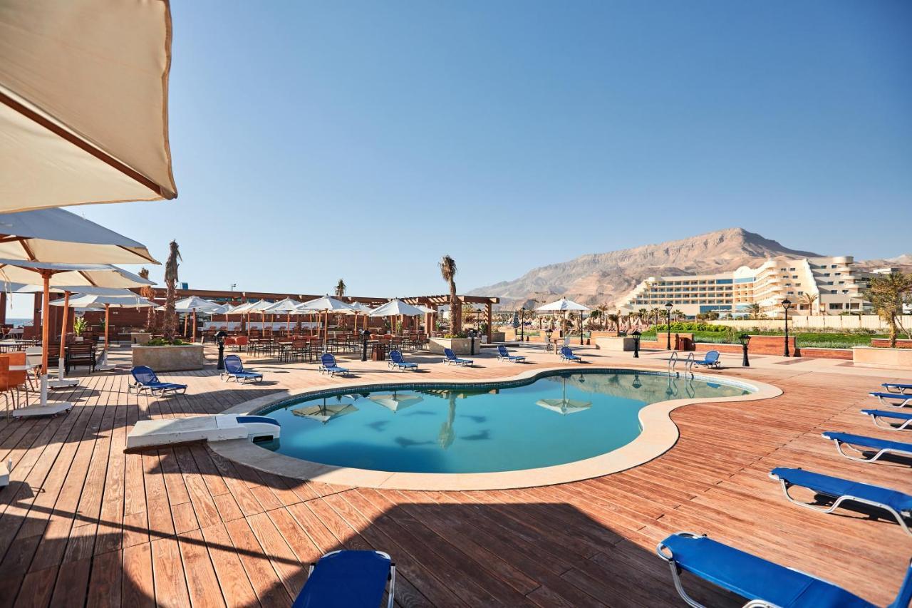 Tolip Resort El Galala Majestic Ain Sukhna Ngoại thất bức ảnh The photo shows a swimming pool area surrounded by sun loungers and umbrellas. The pool has a unique, curvy shape and is situated on a wooden deck. In the background, there are several buildings, likely part of a resort or hotel, and a mountainous la