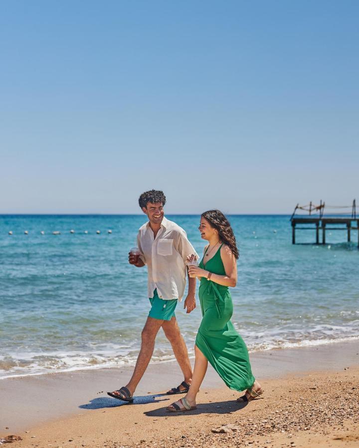 Tolip Resort El Galala Majestic Ain Sukhna Ngoại thất bức ảnh The photo shows a couple walking along a beach. The man is wearing a white short-sleeved shirt and green shorts, while the woman is dressed in a flowing green dress. They both appear to be enjoying the pleasant weather, with the ocean in the backgrou