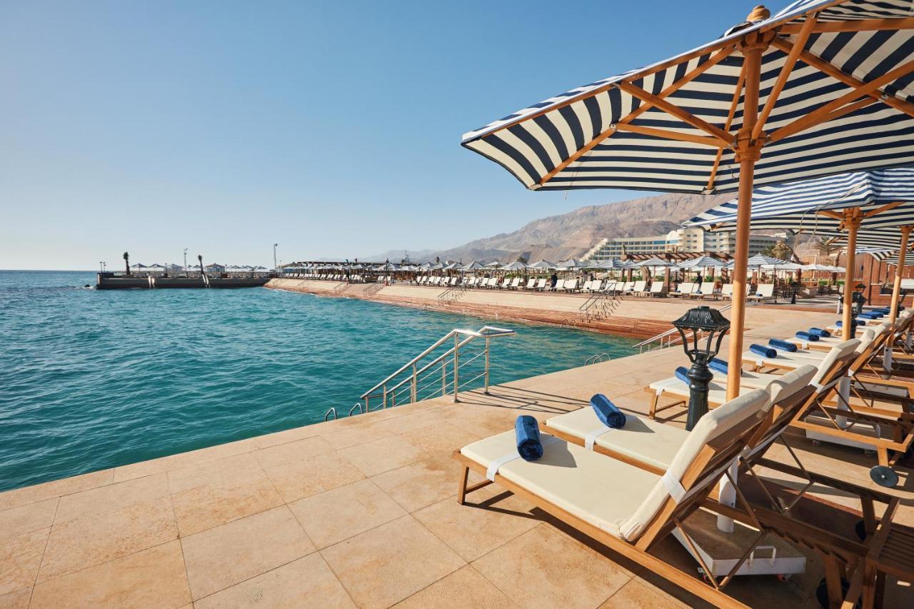 Tolip Resort El Galala Majestic Ain Sukhna Ngoại thất bức ảnh The photo shows a scenic beachside area with clear blue water. In the foreground, there are several sun loungers set up on a tiled surface, with blue towels neatly rolled on each one. The loungers are shaded by striped umbrellas. In the distance, a w