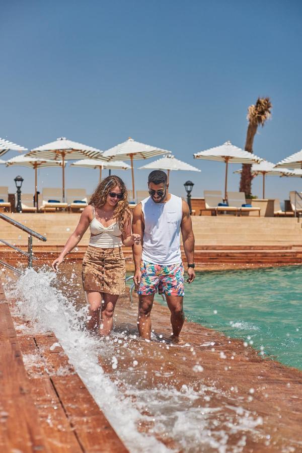 Tolip Resort El Galala Majestic Ain Sukhna Ngoại thất bức ảnh The photo depicts a couple walking along a poolside area. They are enjoying a sunny day near a swimming pool, with splashes of water around them. The woman is wearing a light-colored top and a patterned skirt, while the man is dressed in a sleeveless