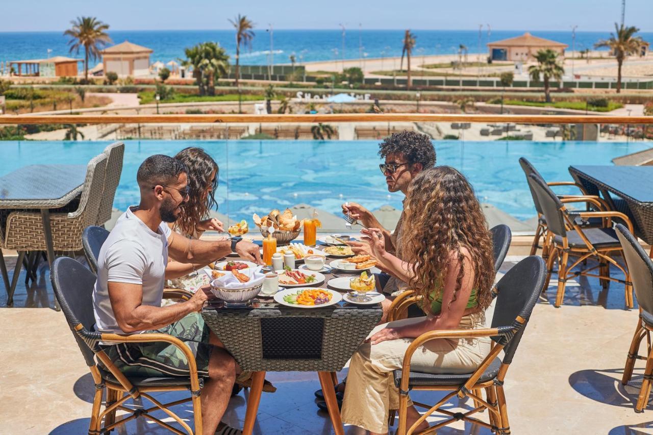 Tolip Resort El Galala Majestic Ain Sukhna Ngoại thất bức ảnh The photo shows a group of five people enjoying a meal together outdoors, likely at a restaurant or cafe. They are seated at a table with a beautiful view of a swimming pool and the sea in the background. The table is filled with various dishes, and 