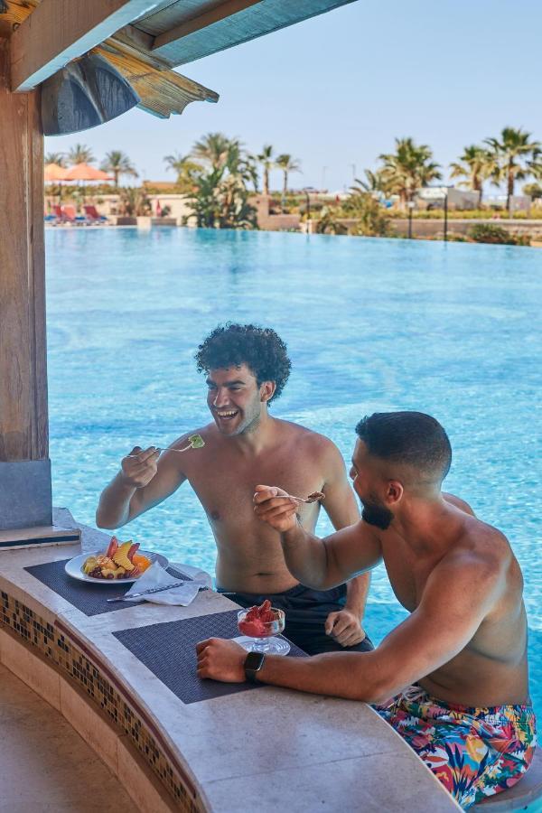 Tolip Resort El Galala Majestic Ain Sukhna Ngoại thất bức ảnh The photo features two young men enjoying a meal together. They are sitting at a poolside table with a clear blue swimming pool in the background. The men are shirtless, suggesting a casual, relaxed atmosphere, likely in a warm environment. One man i