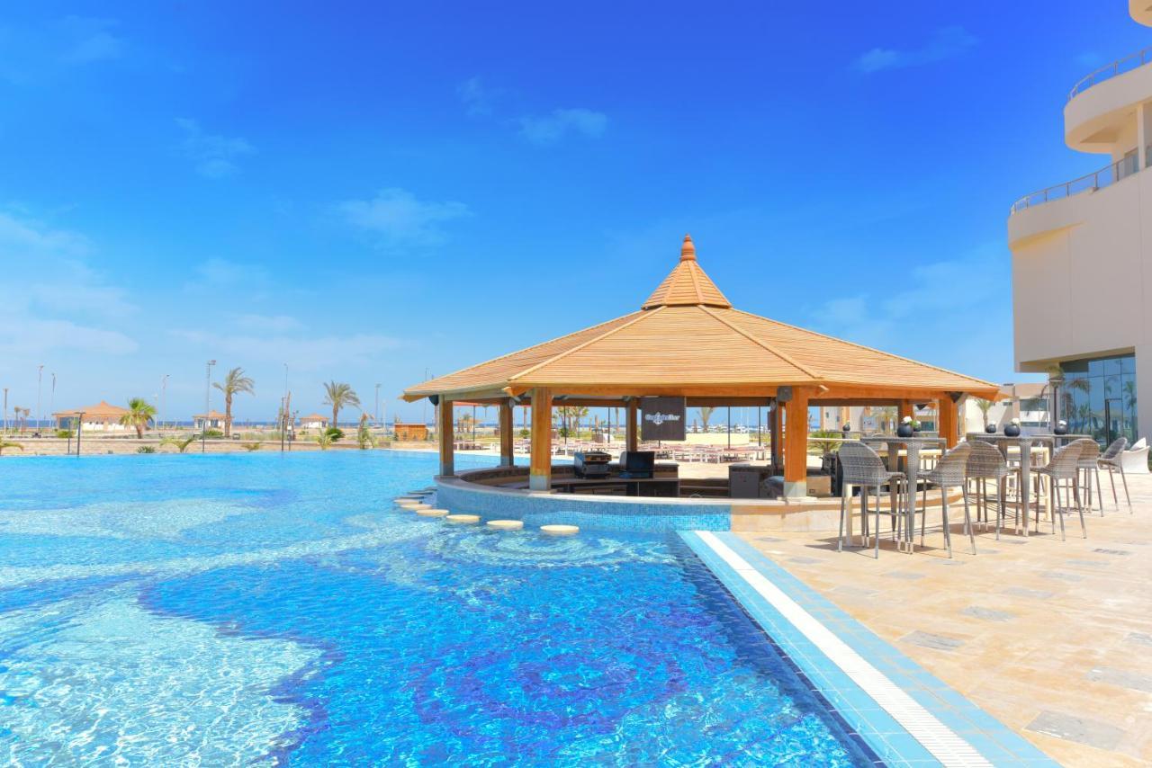 Tolip Resort El Galala Majestic Ain Sukhna Ngoại thất bức ảnh The photo shows a beautiful outdoor setting featuring a large swimming pool with clear blue water. In the center, there is a thatched-roof gazebo, which serves as a shaded seating area. Surrounding the gazebo are lounge chairs and a bar area with hig