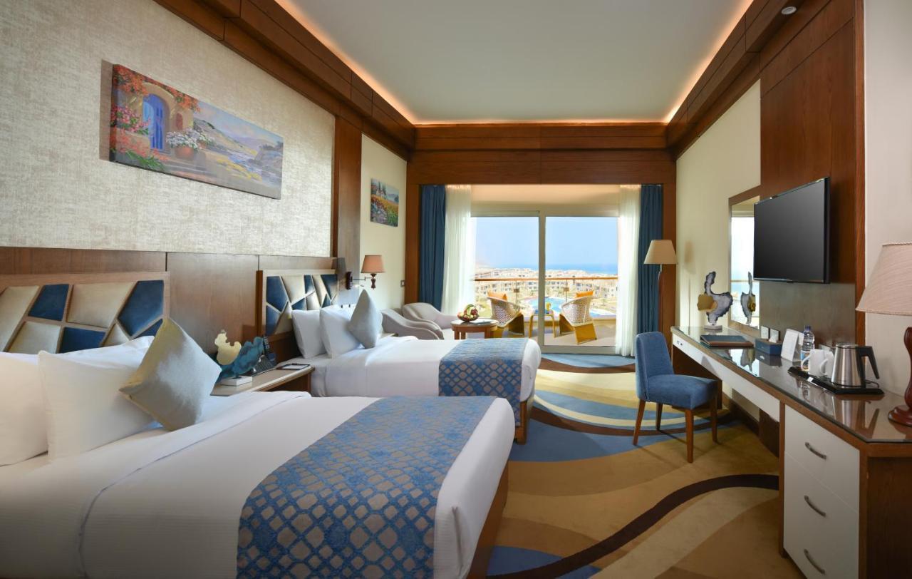 Tolip Resort El Galala Majestic Ain Sukhna Ngoại thất bức ảnh The photo shows a well-decorated hotel room featuring two beds with decorative pillows. The room has wooden accents and modern furnishings, including a desk, a television, and lamps. Large windows flood the space with natural light, offering a view o