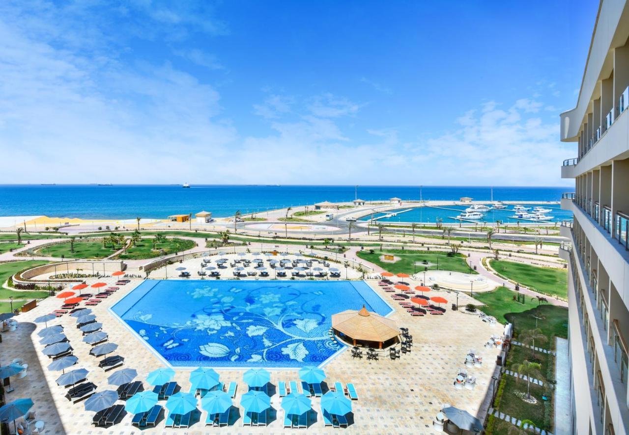 Tolip Resort El Galala Majestic Ain Sukhna Ngoại thất bức ảnh The photo shows a beautiful outdoor view of a resort area featuring a large swimming pool with a decorative underwater design. Surrounding the pool are numerous sun loungers and umbrellas in a variety of colors. In the background, there is a scenic c
