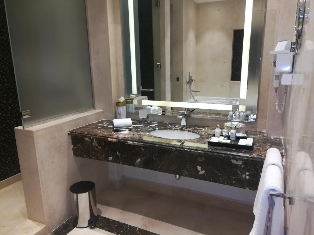 Tolip Resort El Galala Majestic Ain Sukhna Ngoại thất bức ảnh The photo shows a modern bathroom with elegant design features. There is a large vanity made of dark marble with a sink and a set of toiletries neatly arranged on top. A mirror with built-in lighting is mounted above the sink. To the side, there is a