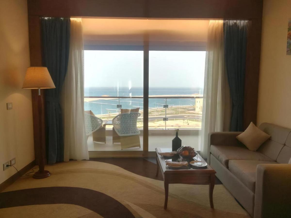 Tolip Resort El Galala Majestic Ain Sukhna Ngoại thất bức ảnh The photo shows a stylish interior of a room with a large window overlooking the sea. The window features sheer and thick curtains and is framed by a balcony with two chairs visible outside. Inside, there is a comfortable-looking sofa, a coffee table