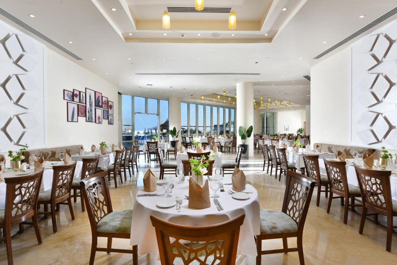 Tolip Resort El Galala Majestic Ain Sukhna Ngoại thất bức ảnh The photo displays a spacious and elegantly designed restaurant interior. It features numerous tables set for dining, with white tablecloths, neatly folded napkins, and decorative centerpieces of flowers. The seating consists of wooden chairs that co