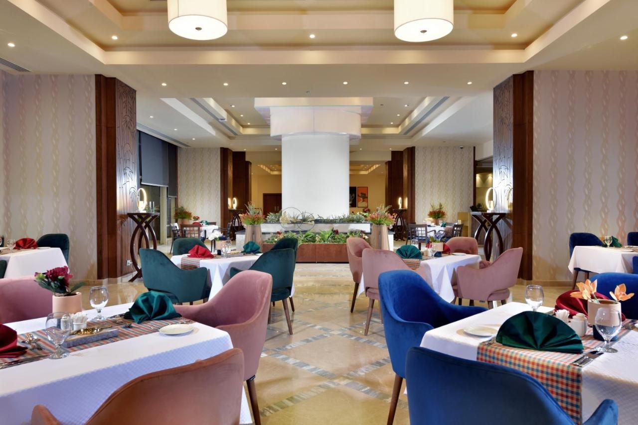 Tolip Resort El Galala Majestic Ain Sukhna Ngoại thất bức ảnh The photo shows a spacious and elegantly designed restaurant interior. The layout includes several round tables dressed in white tablecloths, each set with decorative plates, cutlery, and colorful napkins. The chairs are upholstered in a variety of c